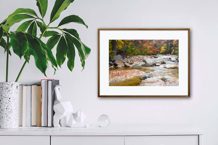 Framed landscape print of a river with autumn foliage on a white wall above books and decor.