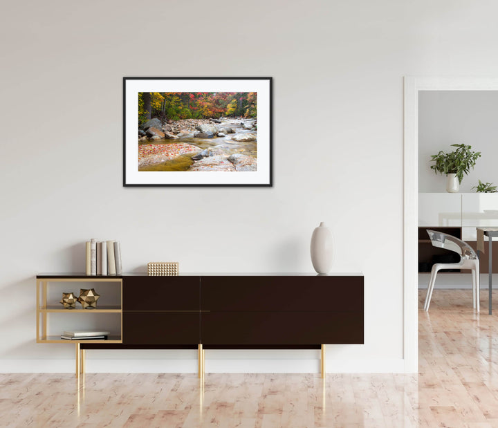 Framed landscape print of a flowing stream with red leaves on a fallen rock and bright autumn trees along the shore. The photograph is on a wall above a dark piece of furniture in a modern room.