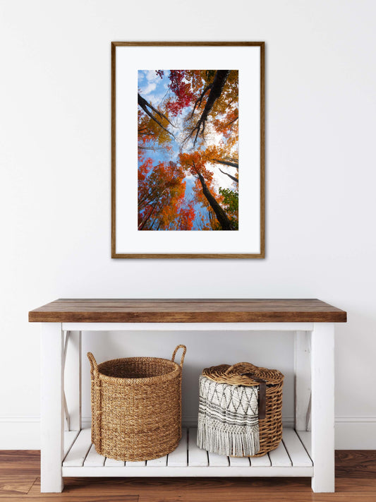 Framed artwork of autumn trees above a wooden table with baskets on a white wall.
