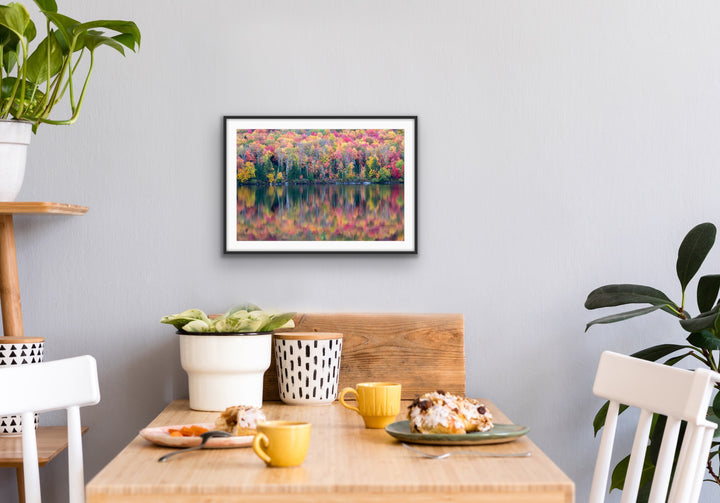 Dining room with a table set for breakfast, plants, and a framed landscape print on the wall. The landscape photograph on the wall is a brightly colorful autumn forest reflected in a calm lake. 
