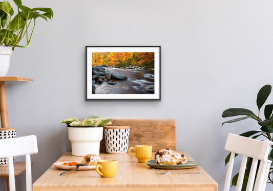 Framed photograph of a autumn landscape with a flowing stream and colorful trees along the shore, on a wall above a dining table with food and drinks.